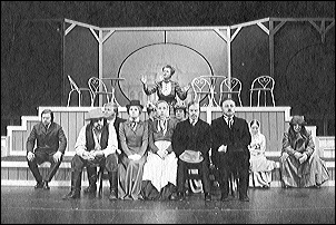 Kuva Minna -näytelmästä. Näyttelijät istuvat penkeillä näyttämöllä. Taustalla näkyvät lavasteet.