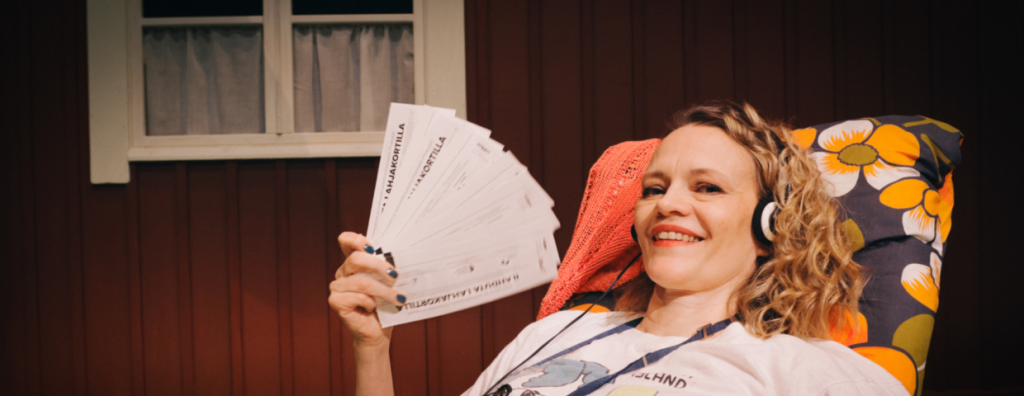 Nainen aurinkotuolissa rennosti hymyillen, pitelee kädessään teatterin pääsylippuja viuhkana.